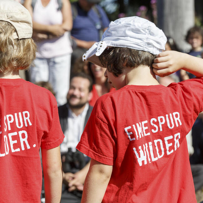 Von hinten stehen Kinder vor eine Gruppe von Menschen. Auf ihren T-Shirt steht "Eine Spur wilder".