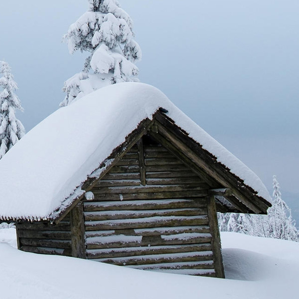 Eine kleine Holzhütte mit dick zugeschneitem Dach. Auch die restliche Landschaft ist mit hihem Schnee bedeckt.