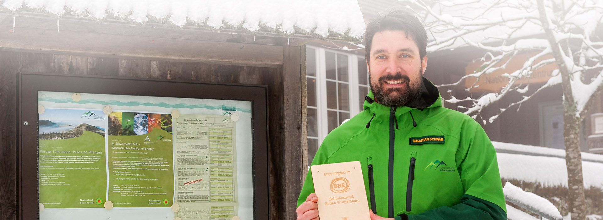 Sebastian Schwab vor dem verschneiten Infozentrum des Nationalparks. Er hält eine Holztafel mit der Aufschrift „Ehrenmitglied im BNE-Schulnetzwerk Baden-Württemberg“ in die Kamera. Links neben ihm ist ein Schaukasten des Nationalparks mit Veranstaltungsaushängen zu sehen.