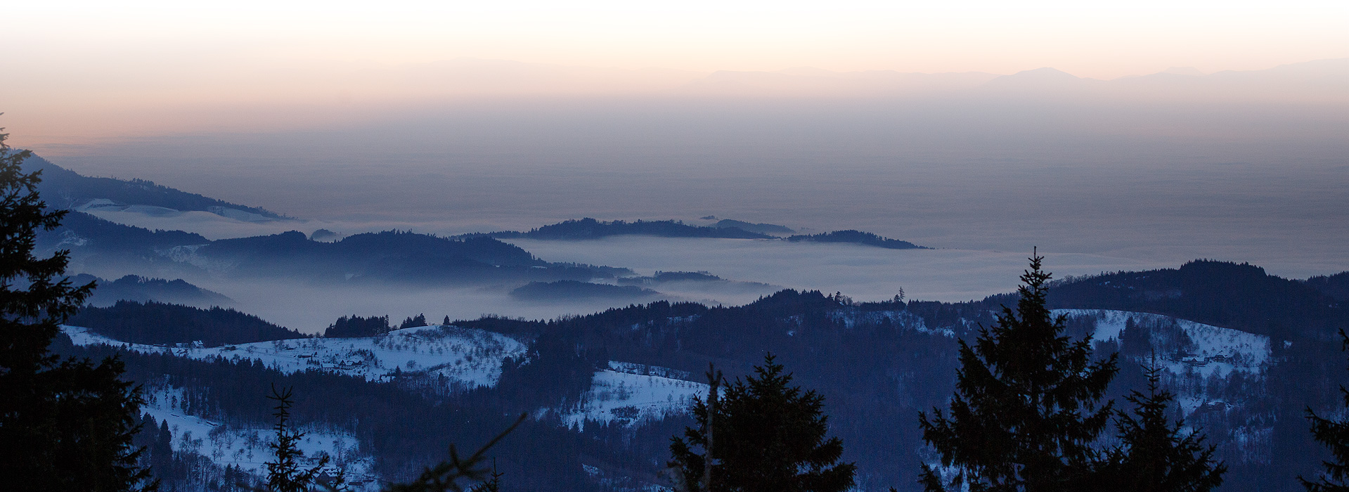 Ausblick über mehrere mit etwas Schnee bedeckten Berggipfel. Die Rheinebene dahinter verschwindet im Nebel, der auch durch die Täler zieht. 