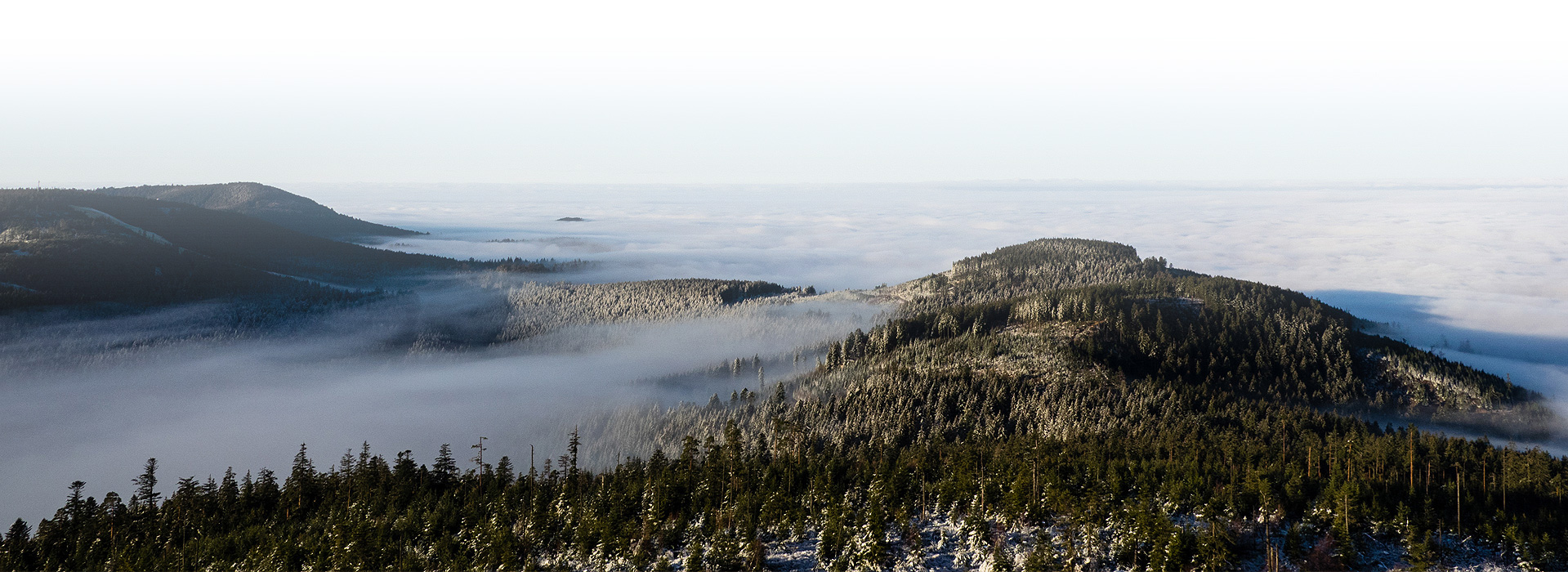 Ausblick über schneebestäubte Berge. Zwischen den Bergen hängt Nebel, die Ebene hinter den Bergen verschwindet in einem Wolkenmeer.