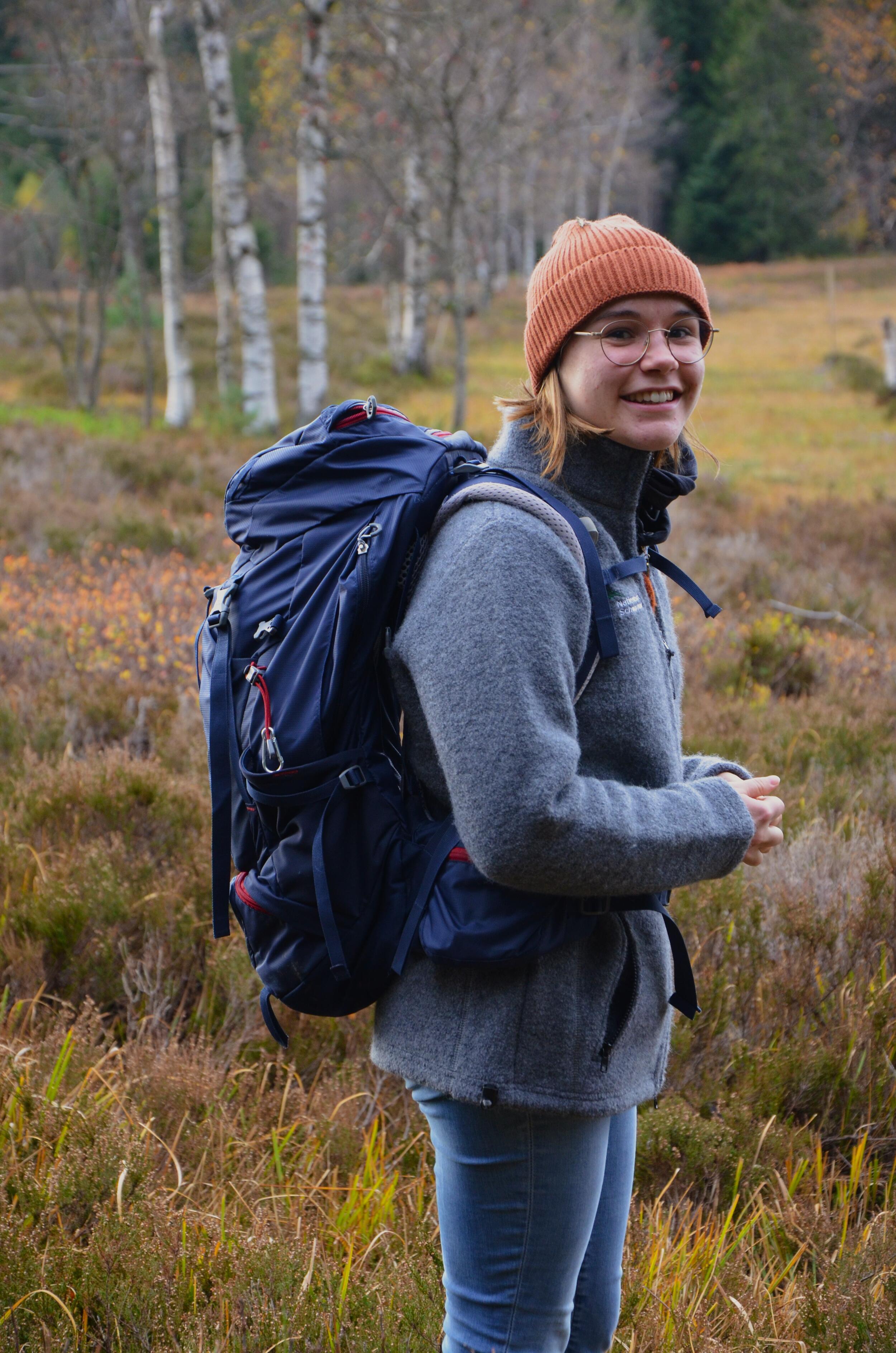 Die Freiwilligendienstleistende Elsa steht in der Natur vor einer Moorlandschaft mit Birken im Hintergrund und trägt eine Mütze