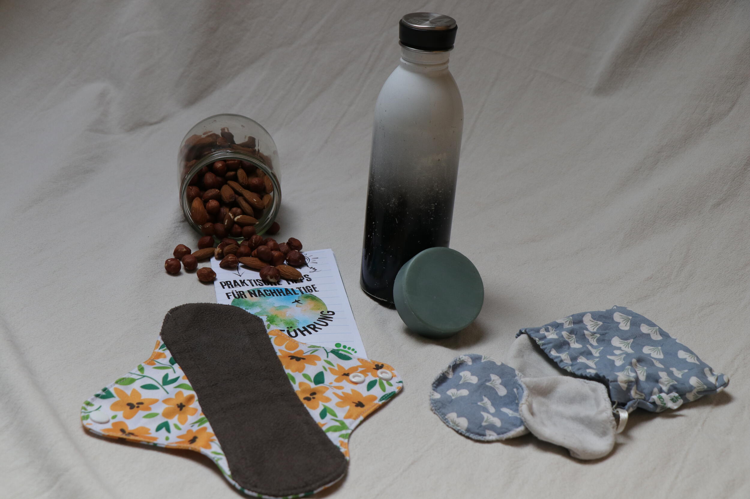 Nüsse in einem Glas, eine wiederbefüllbare Trinkflasche, eine wiederverwendbare Binde und andere wiederverwendbare Gegenstände liegen auf einem Tuch