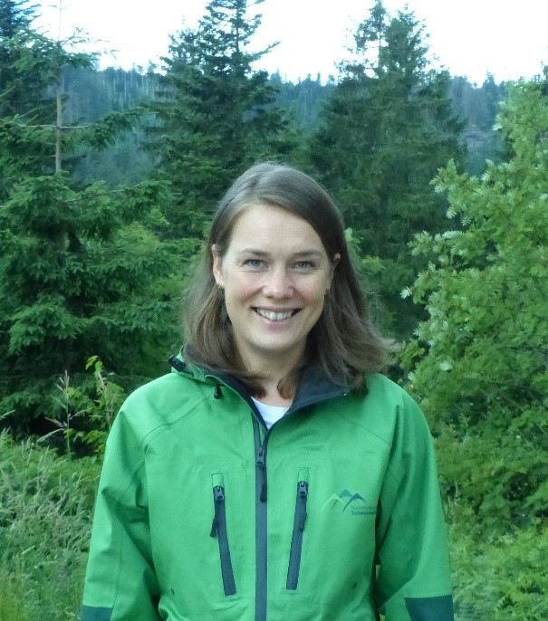 Eine Frau in grüner Jacke steht vor einem Wald.