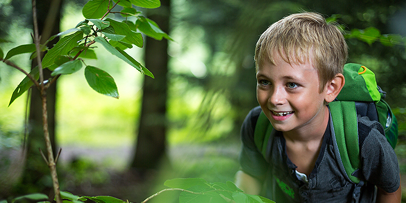 Junge mit Rucksack im Wald, schaut neugierig lachend.