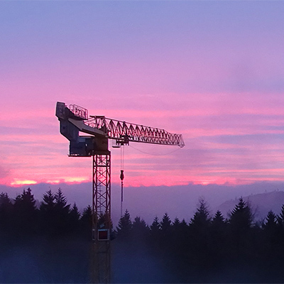 Ein Baukran vor einem vom Sonnenuntergang blau und  rosa gefärbten Himmel und einer dunklen Siluette von Nadelbäumen.