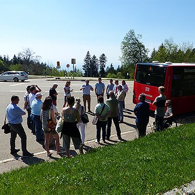 Eine Gruppe steht im Kreis am Rand eines Parkplatzes. Ein Teilnehmer sitzt im Rollstuhl. Rechts ist ein Bus zu sehen.