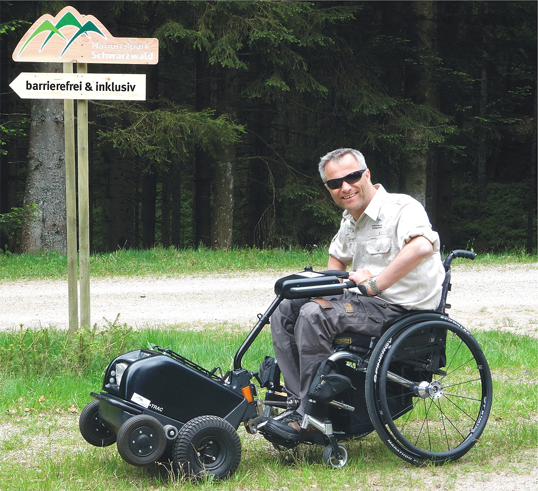 Ein Rollstuhlfahrer vor einem Nationalparkschild. Vor seinem Rollstuhl steht eine kleine Zugmaschine.