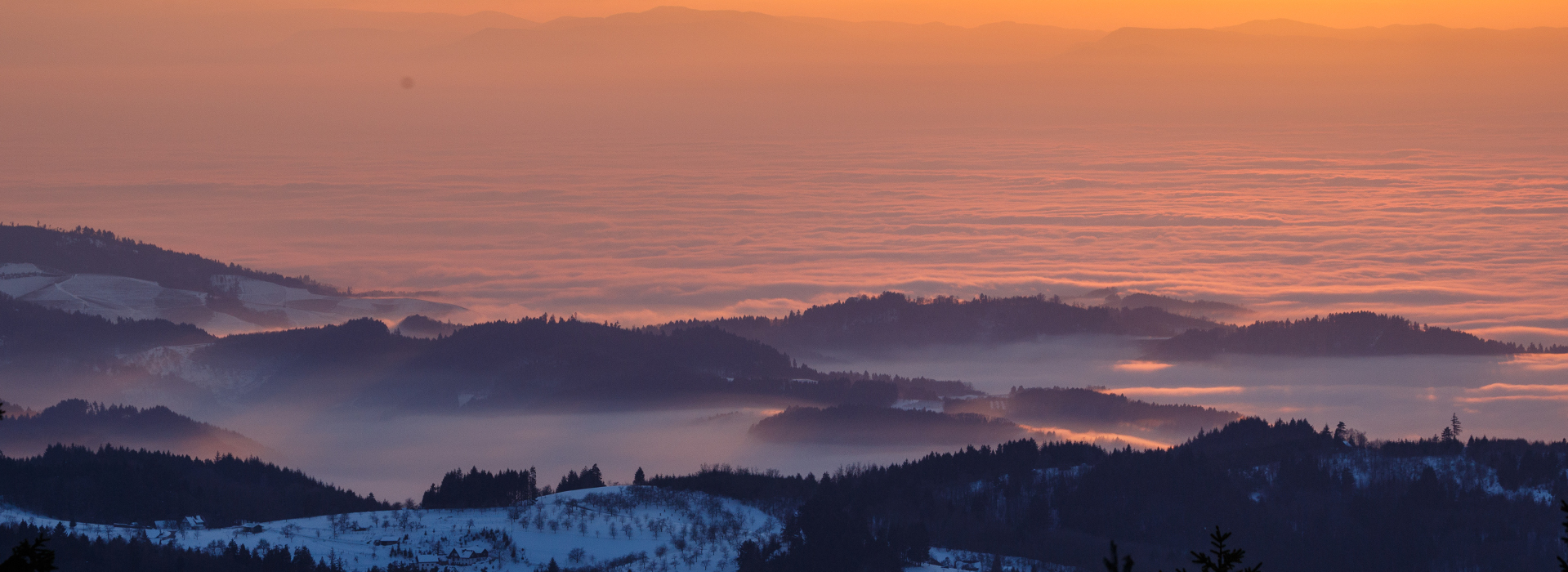 Ausblick über mehrere leicht verschneite Berggipfel in die mit dicken Wolken gefüllte Rheinebene. Zwischen den Berggipfeln hängen Nebelschleier, die Wolken werden vom Sonnenuntergang rötlich verfärbt.  