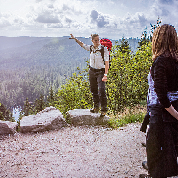 Eine Rangerin steht auf einigen großen Steinen und erklärt einer Besucherin etwas über die Aussicht hinter ihr.