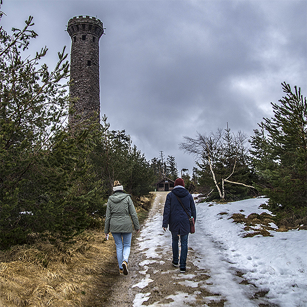 Auf der Bergkuppe steht ein Steinturm. Links davon führt ein Weg auf eine Schutzhütte zu. Zwei Menschen laufen auf dem Weg. Es liegt etwas Schnee.