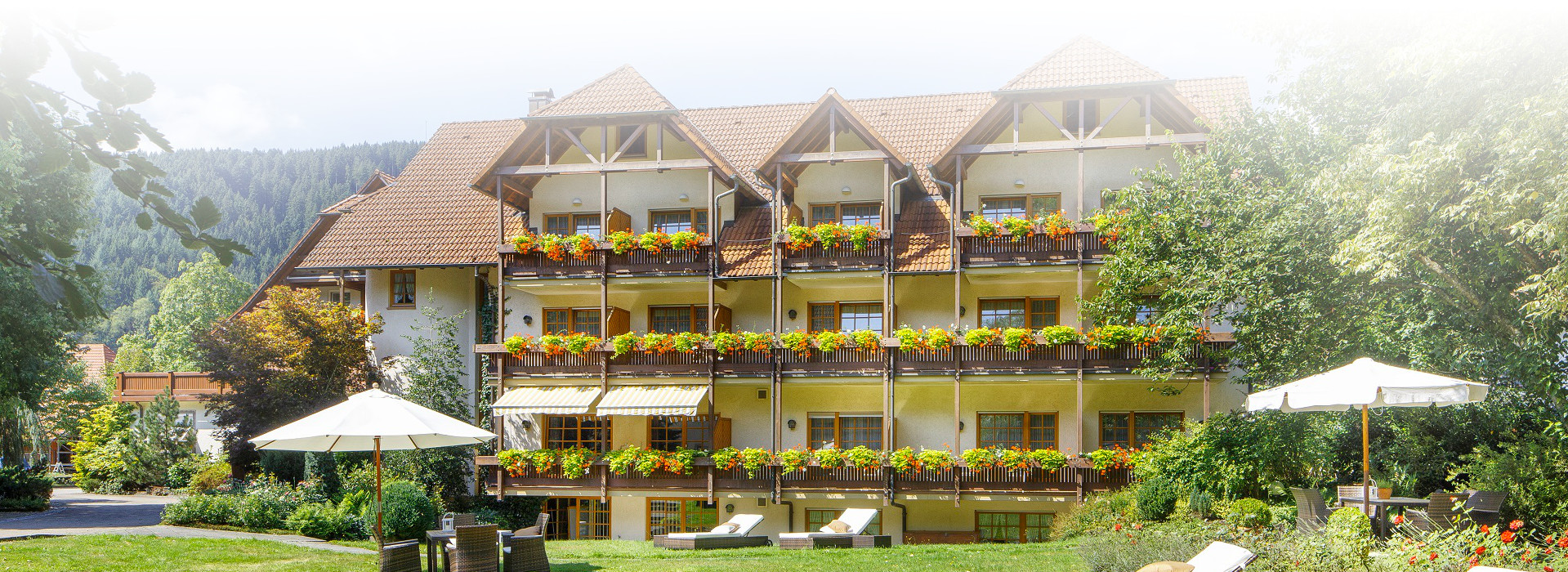Ein Hotel mit vielen Balkonen. An jedem Balkon sind Blumenkästen befestigt. Im Garten vor dem Hotel stehen Liegen und verteilte Sitzgruppen mit Sonnenschirmen und Tischen.