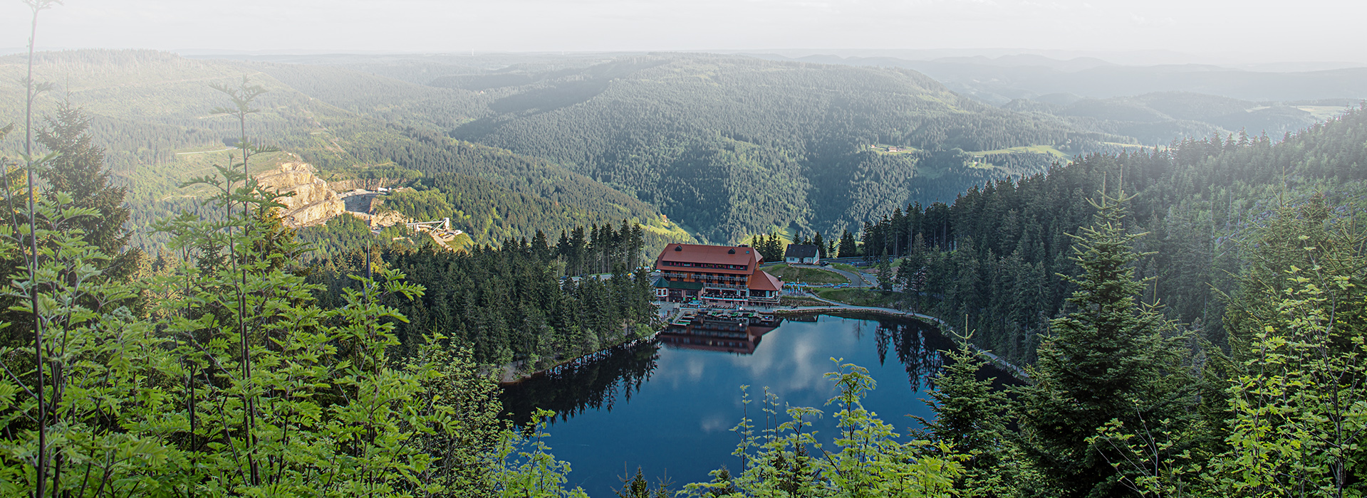 Blick auf das Berghotel Mummelsee. Zwischen dichtem Nadelwald liegt der runde Mummelsee. An seinem Ufer steht ein großes Haus. Im Hintergrund sind Berghänge zu sehen.
