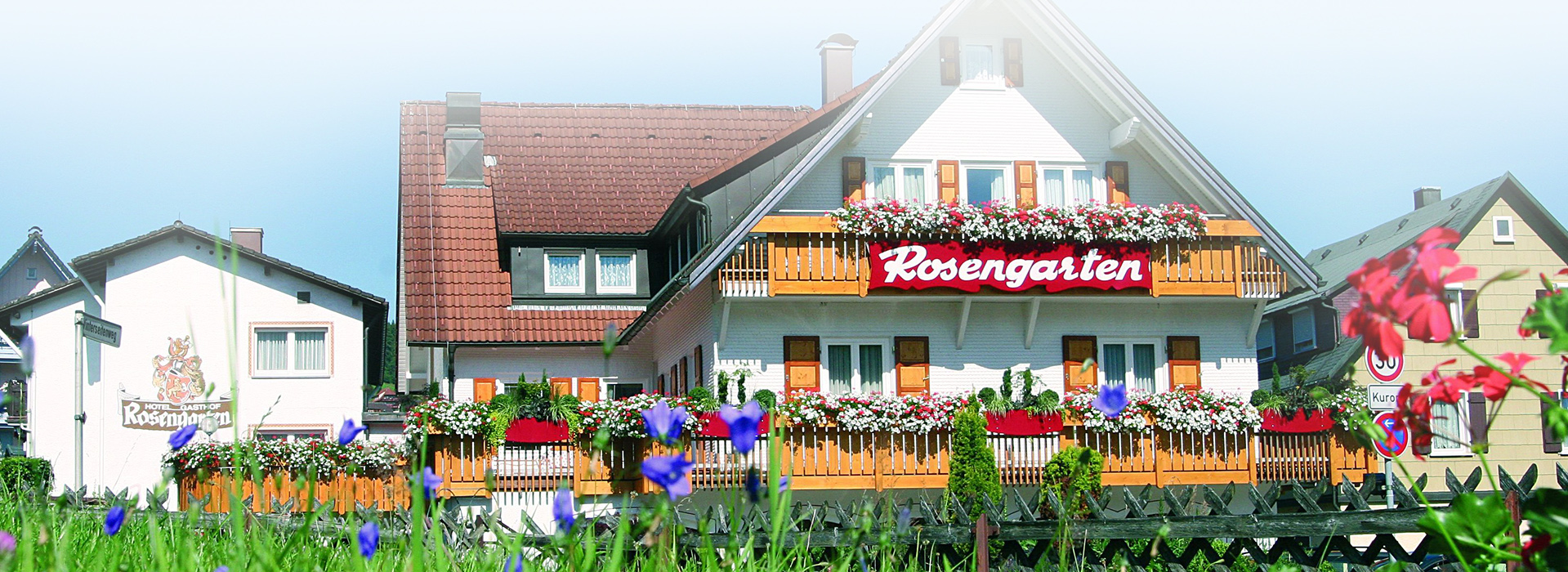 Ein großes Haus mit mehreren großen Balkonen, an denen Blumenkästen hängen. An einem Balkon hängt ein rotes Schild mit der Aufschrift "Rosengarten". Die Fassade besteht aus weißen Schindeln. Neben dem Haus stehen noch weitere.