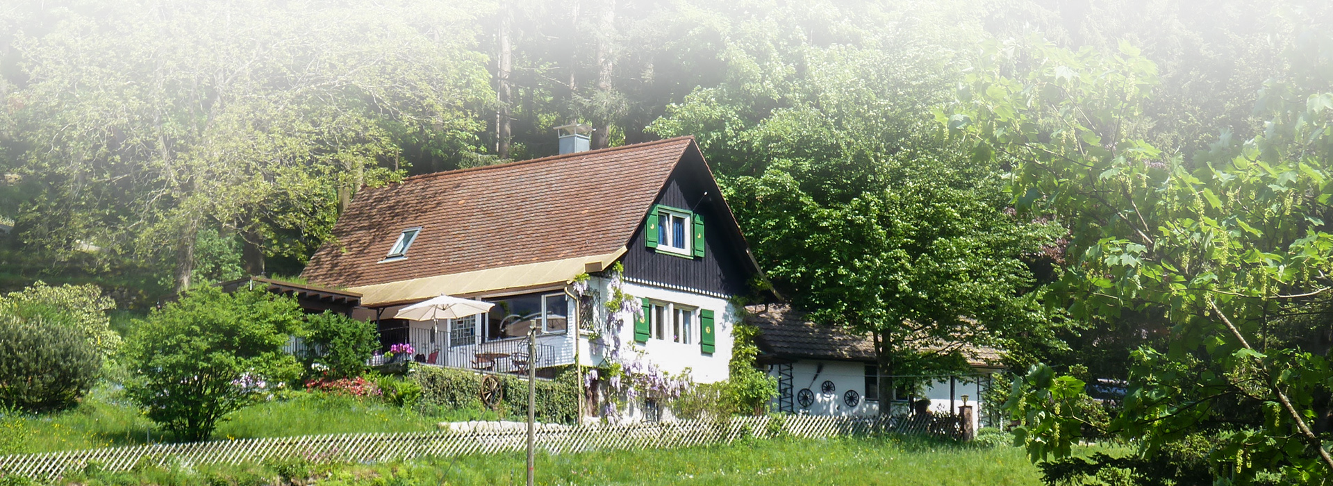 Ein großes, mehrstöckiges Haus mit grünen Fensterläden. An der Seite ist ein niederiger Anbau  zu erkennen. Am Haus ist auch ein großer Garten, der von einem Zaun eingefasst wird. Hinter dem Haus beginnt der Wald.