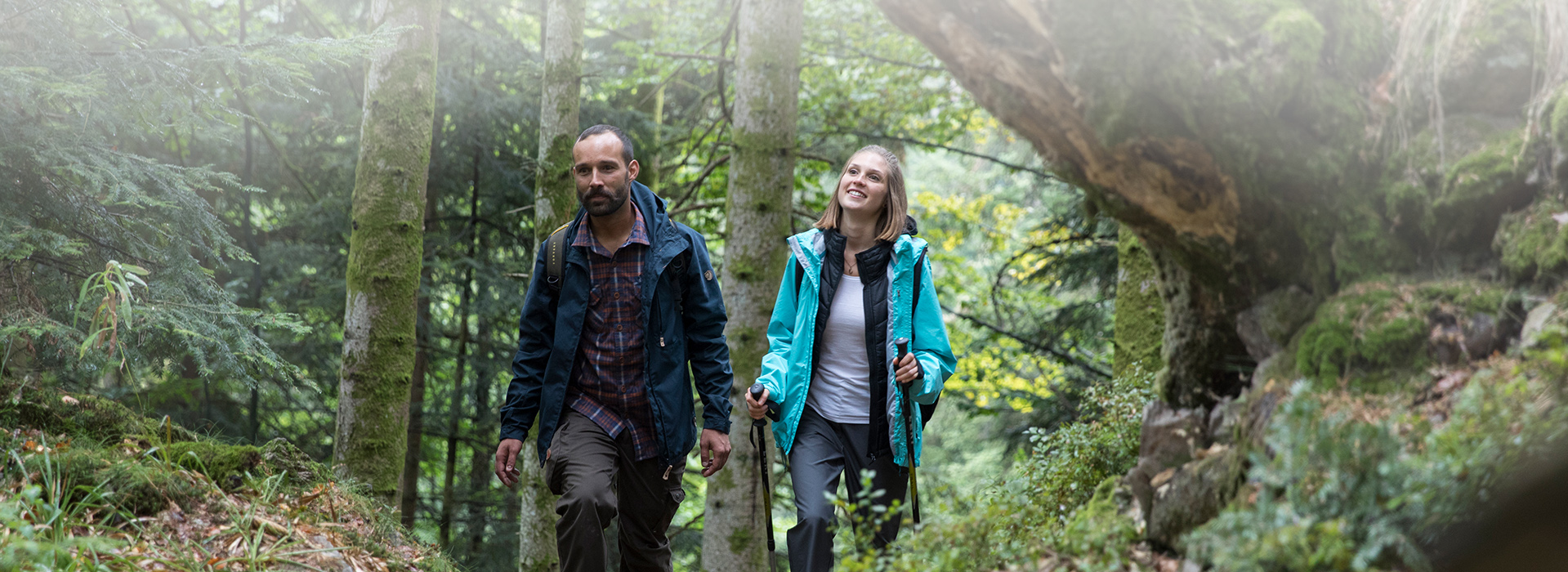 Ein Mann und eine Frau wandern im Wald. Im Hintergrund stehen Bäume, links ragt ein Fels ins Bild. 