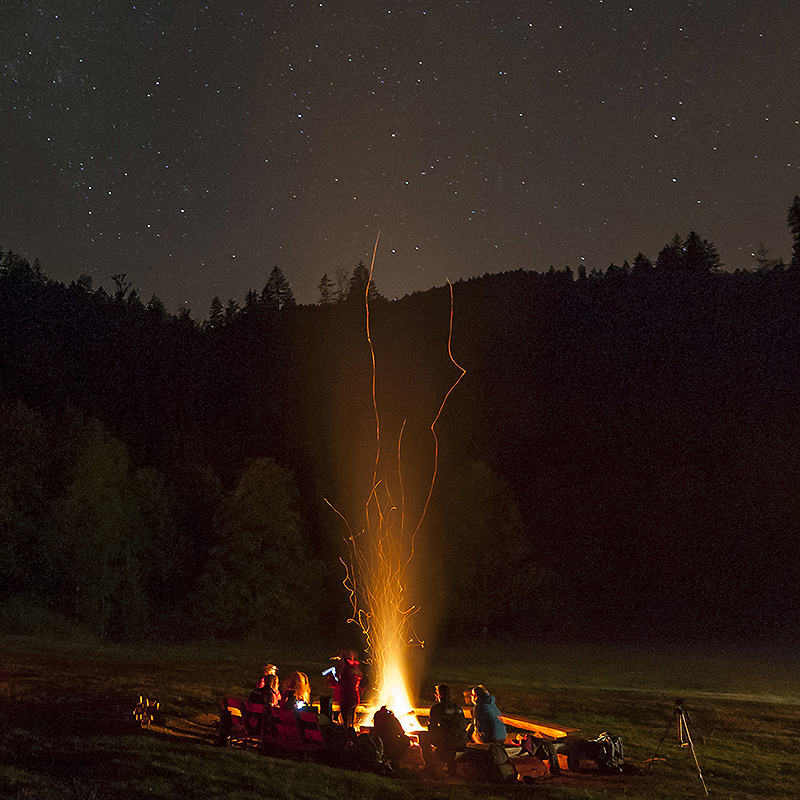 Eine Gruppe Leute sitzt im Kreis um ein Lagerfeuer. Die Funken des Feuers fliegen sehr hoch, am Himmel sieht man Sterne.