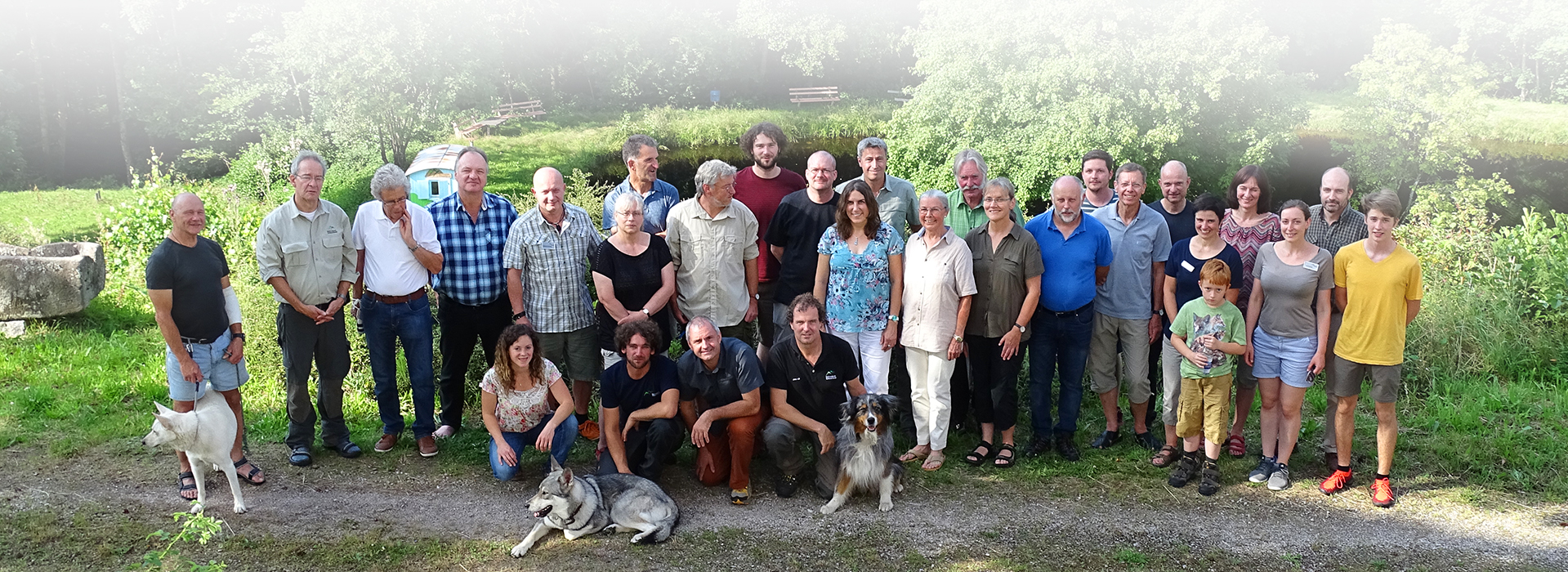 Größere Gruppe Ehrenamtlicher Rangerinnen und Ranger in sommerlicher Kleidung mit See und Bäumen im Hintergrund.