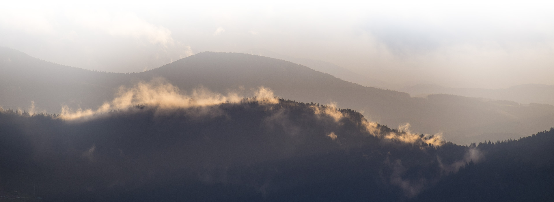 Von einem dunklen Berghang steigt Nebel auf. Der Nebel wird angeleuchtet, im Hintergrund sind weitere Berg zu erahnen.