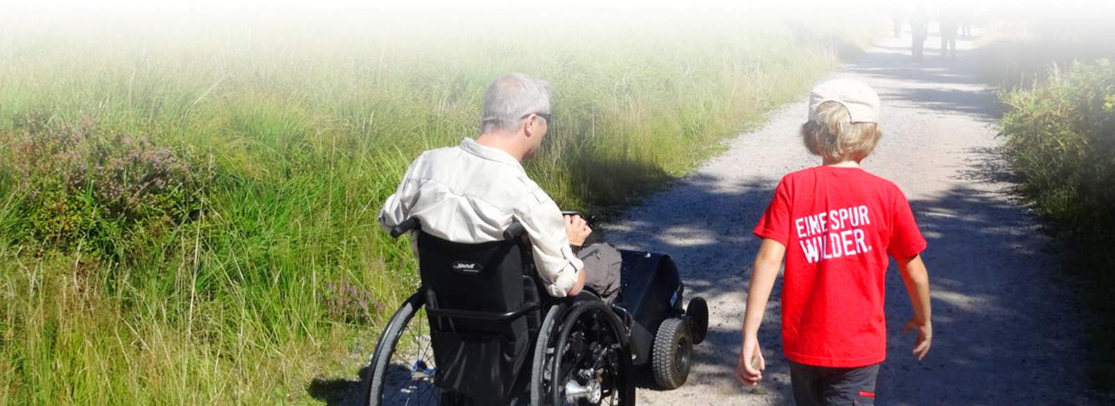 Ein Mann im Rollstuhl mit einer Zugmaschine rollt neben einem Jungen über einen geschotterten Weg.