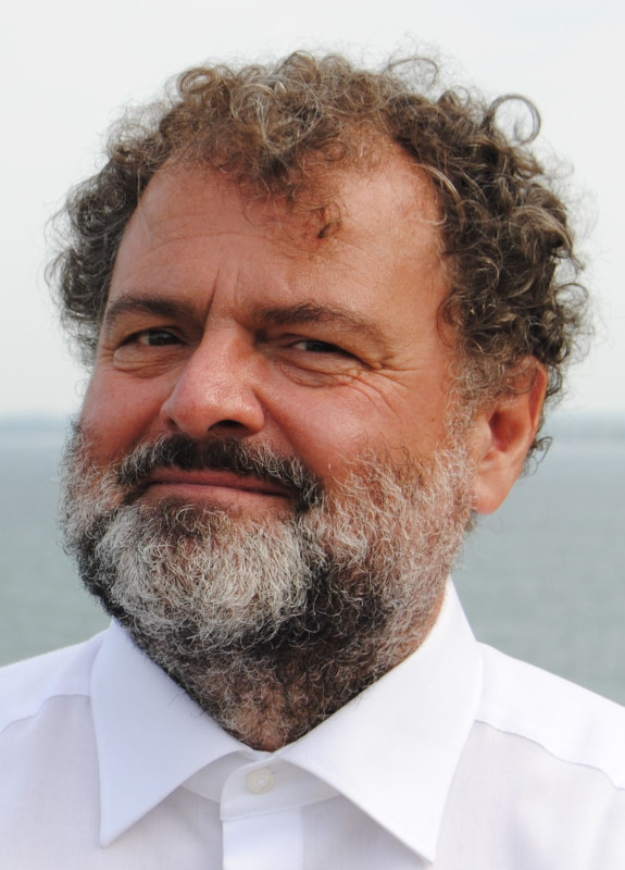 Porträtfoto eines älteren, lachenden Mannes. Er hat krause, mit grau durchzogene dunkelblonde Haare und einen grauen Vollbart.