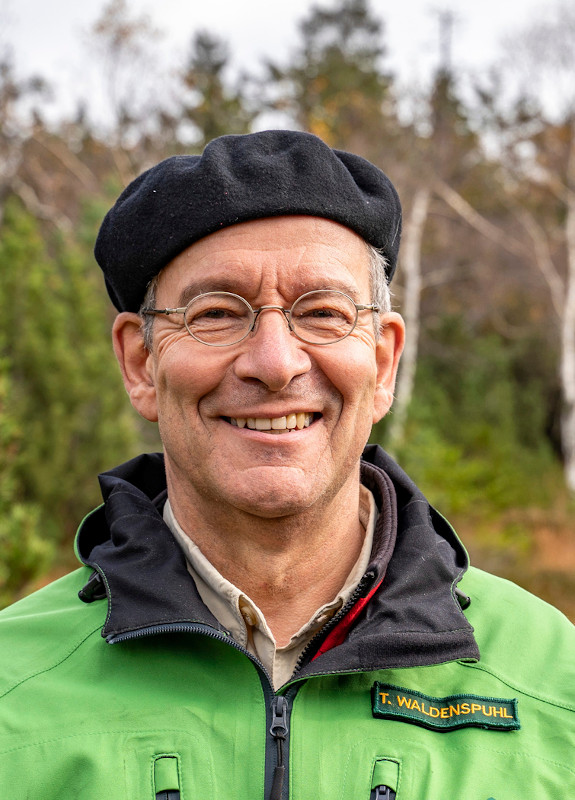 Porträtbild eines älteren Mannes mit dünner Metallrandbrille und blauer Baskenmütze auf grauen Haaren. Er steht vor der Kulisse eines Nadelwaldes und trägt eine grüne Outdoorjacke.