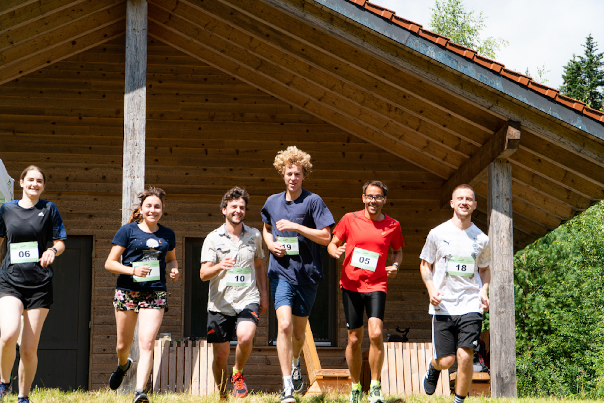Auf dem Bild sind sportlich und kurz gekleidete Läufer zu sehen, mit Nummernschildern auf dem T-Shirt vor einer Holzhütte, die offenbar gut gelaunt sind.