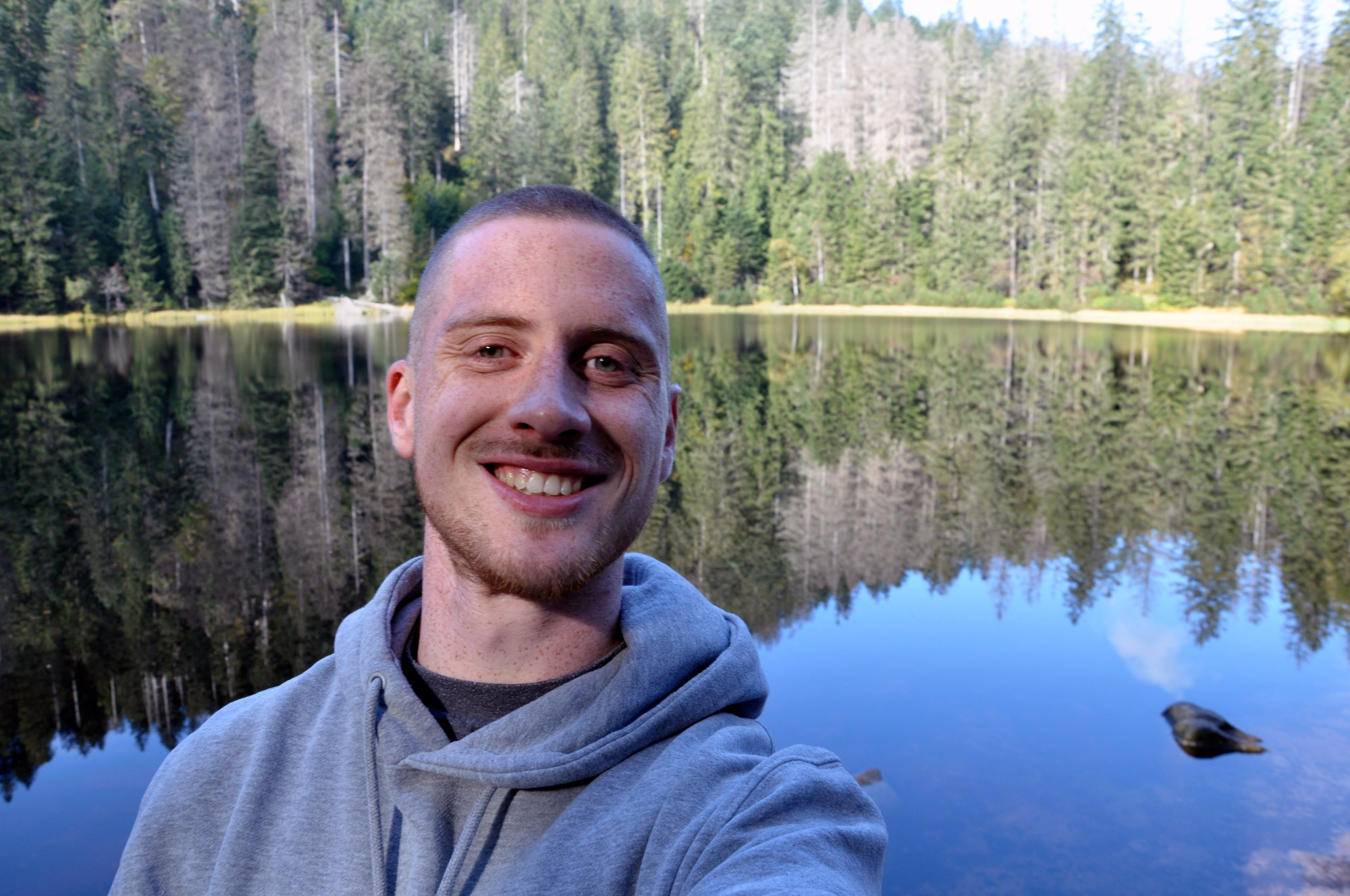 Selfie von Blogger Oliver vor dem Wilden See: Im Hintergrund der ruhig daliegende See mit der Umrahmung durch dichten Nadelwald. Davor ein in die Kamera grinsender junger Mann mit einem grauen Kapuzenpullover.