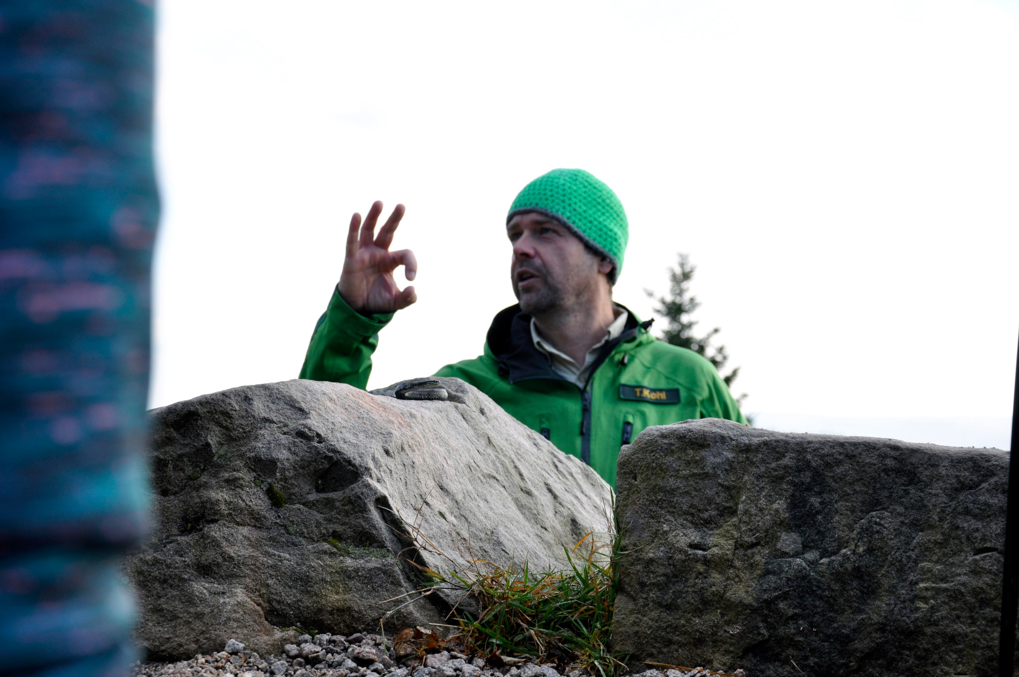 Im Vordergrund sind große Steinblöcke, dahinter steht ein Ranger in grüner Outdoorkleidung und bunt gehäkelter Mütze und zeigt mit seiner rechten Hand eine Art OK-Symbol an.