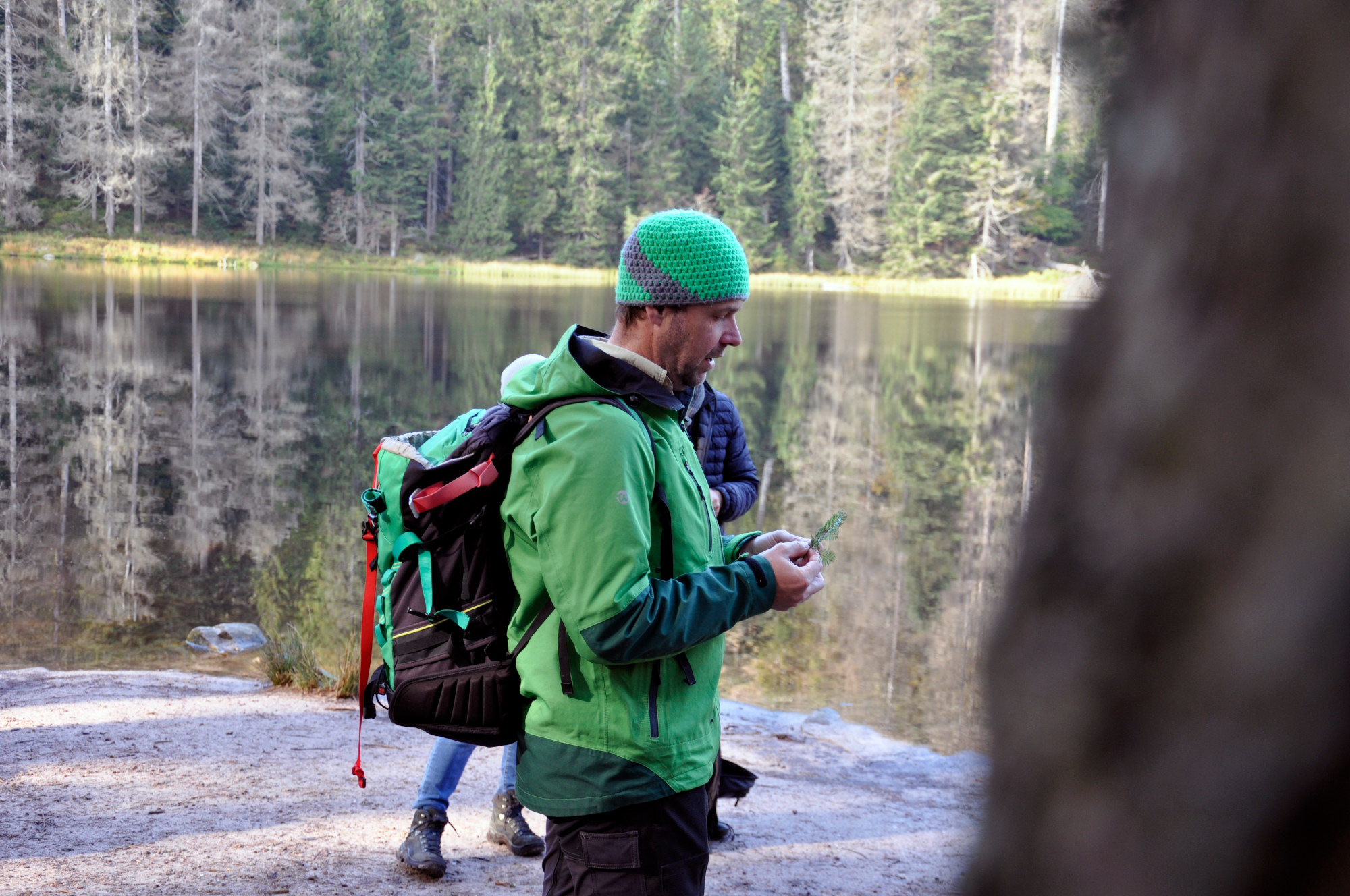 Ranger mit bunt gehäkelter Mütze und herbstlich warmer grüner Outdoorkleidung, der am Ufer eines Sees steht und etwas erklärt.