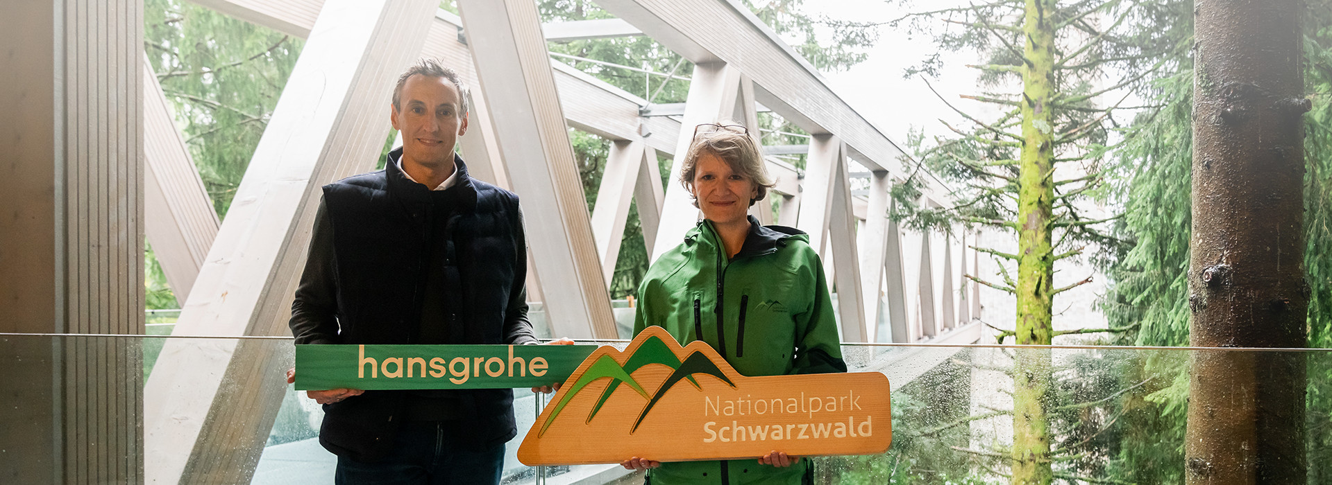 Ein Mann in schwarzer Kleidung (Hans Jürgen Kalmbach) und eine Frau in grüner Jacke (stellvertretende Nationalparkchefin Britta Böhr) halten nebeneinander ihre Logos in die Kamera. Im Hintergrund die Aussichtsbrücke des neuen Nationalparkzentrums. Foto: Hansgrohe SE / retec