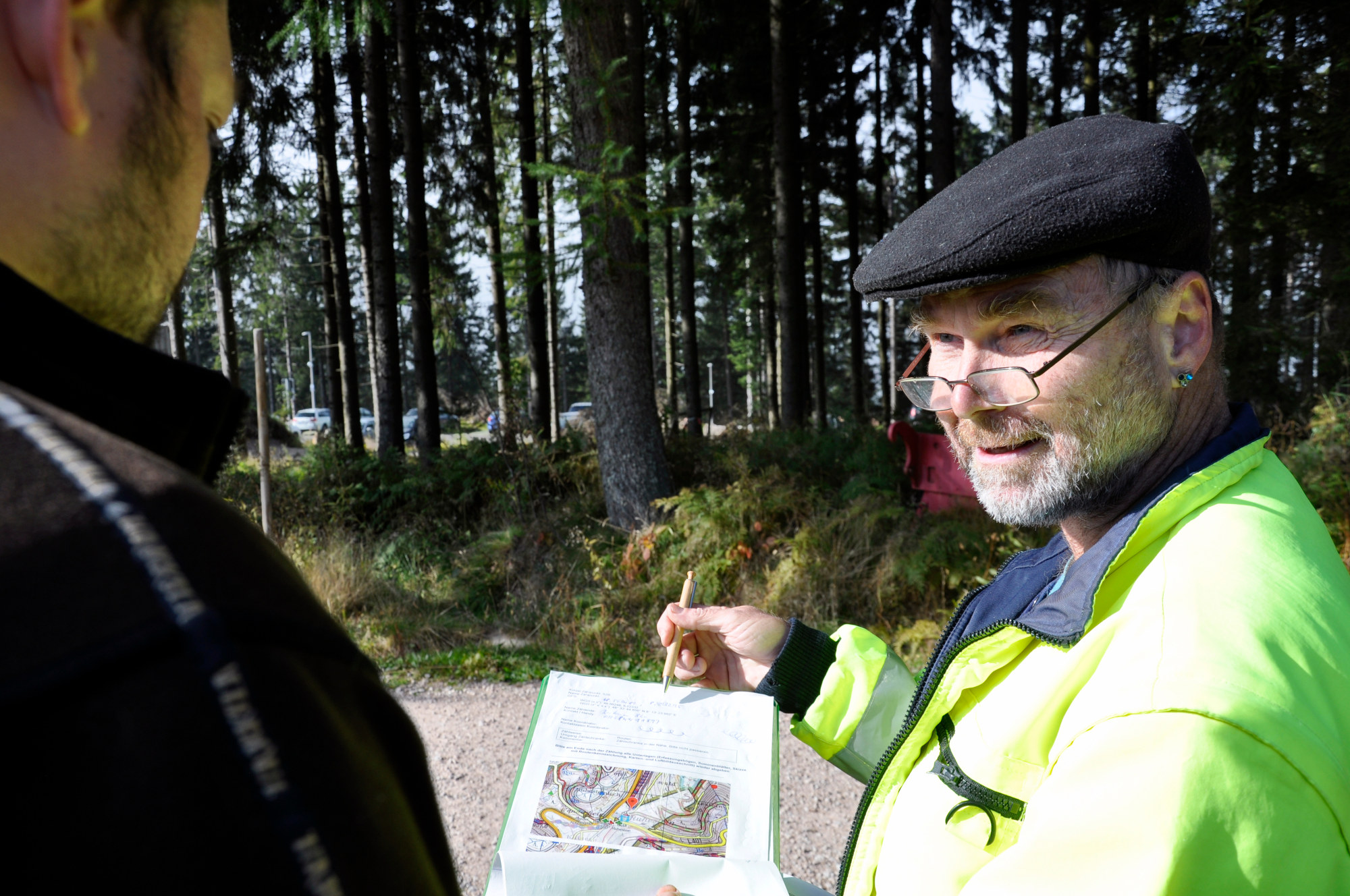 Ein Helfer erklärt einem anderen Mann etwas © Oliver Gewald/ Nationalpark Schwarzwald Ein älterer Herr mit dunkler Schiebermütze, Lesebrille und grauem Vollbart, der etwas erklärt.