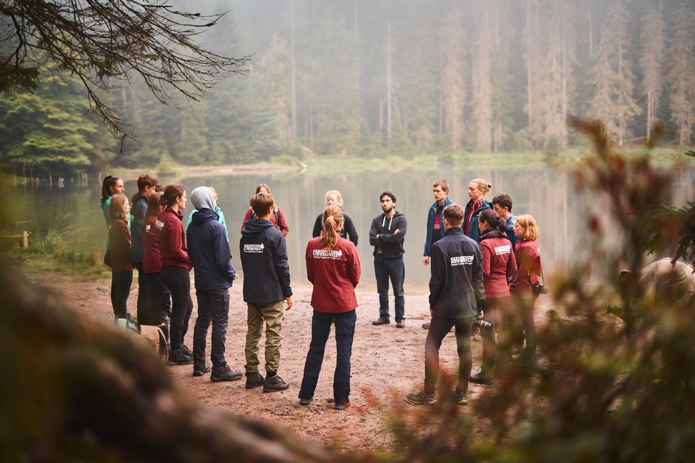 Gruppe junger Leute in blauen und roten Jacken, die in einem Kreis an einem Seeufer stehen.