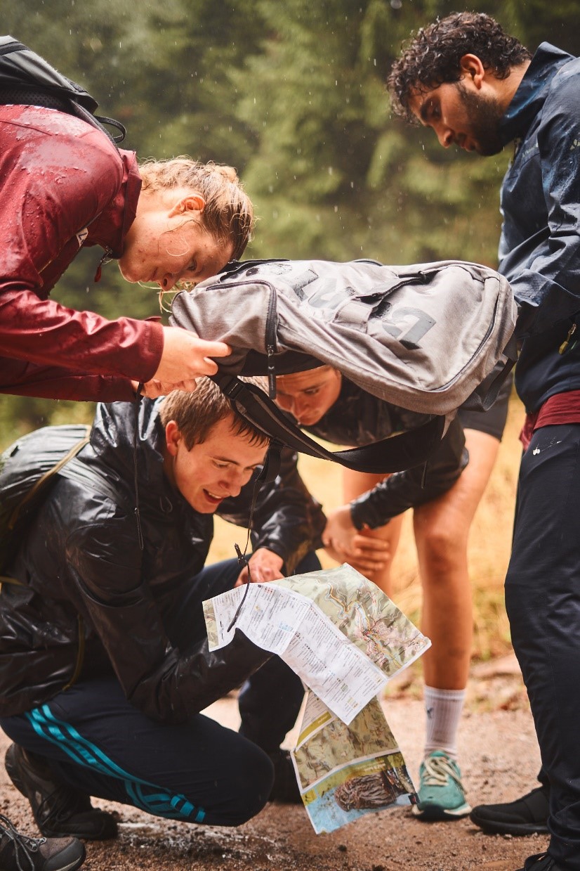 Vier junge Leute in Wanderkleidung versuchen sich gleichzeitig vor dem Regen zu schützen und in eine Karte zu schauen.