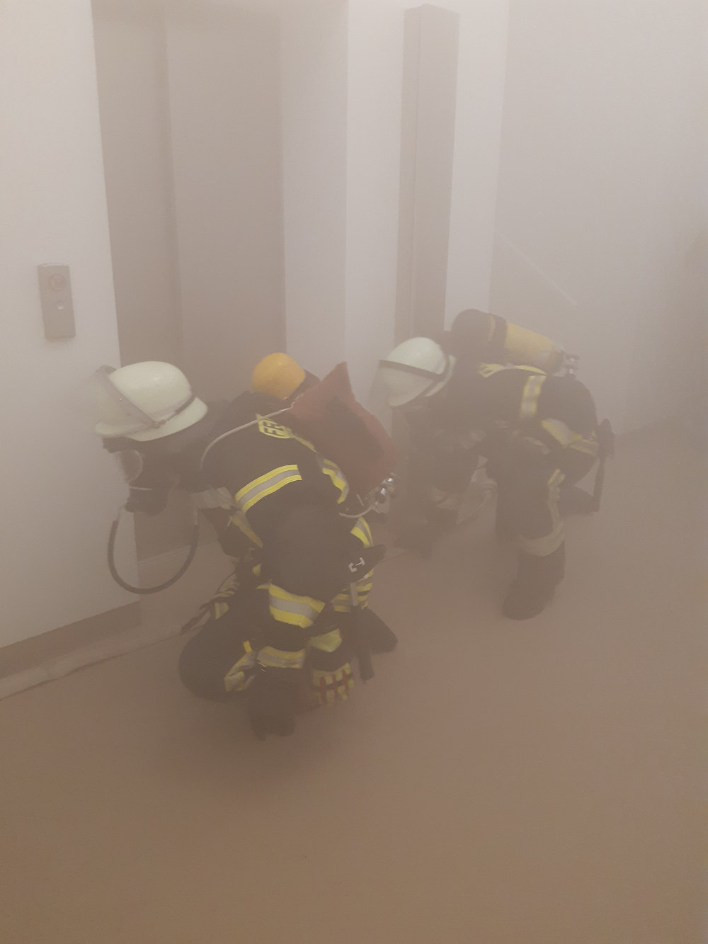 Zwei Feuerwehrleute in geduckter Halteung vor einem geschlossenen Fahrstuhl. Der Raum ist mit hellem Rauch vernebelt.