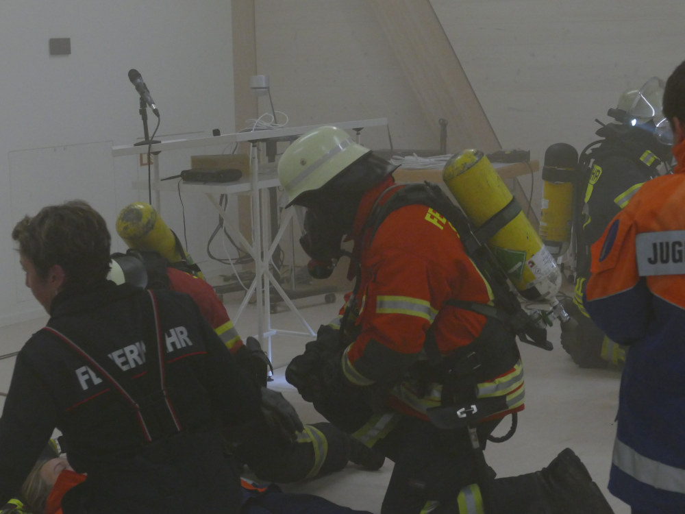 Ein Feuerwehrmann oder eine Feuerwehrfrau mit Atemschutzgerät kniet vor einer verletzten Person. 