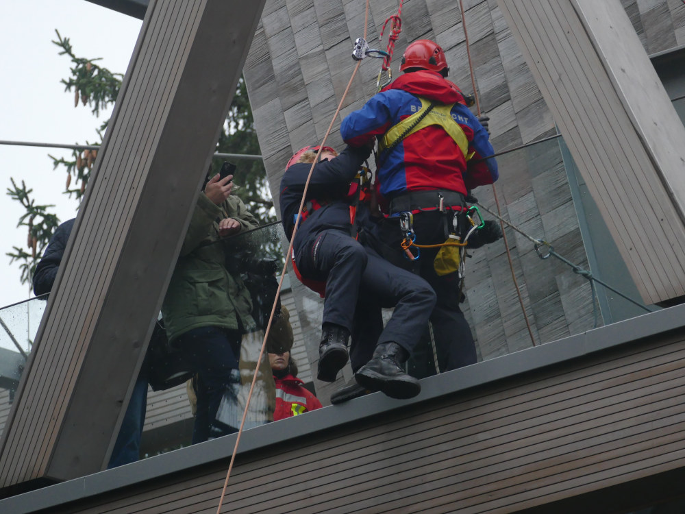 Eine Rettungskraft der Bergwacht steht auf einem metallenen Gittersteg vor einem holzverkleideten Turm und scheint gerade eine verletzte Person in einem Klettergurt an einer Abseilleine zu sichern.