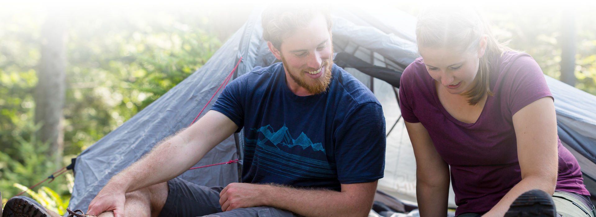 Ein Mann und eine Frau, beide zwischen 20 und 30 Jahre alt, sitzen in sommerlicher Wanderkleidung vor einem kleinen Zelt und schauen in eine Wanderkarte, die zwischen ihnen ausgebreitet auf dem Boden liegt.