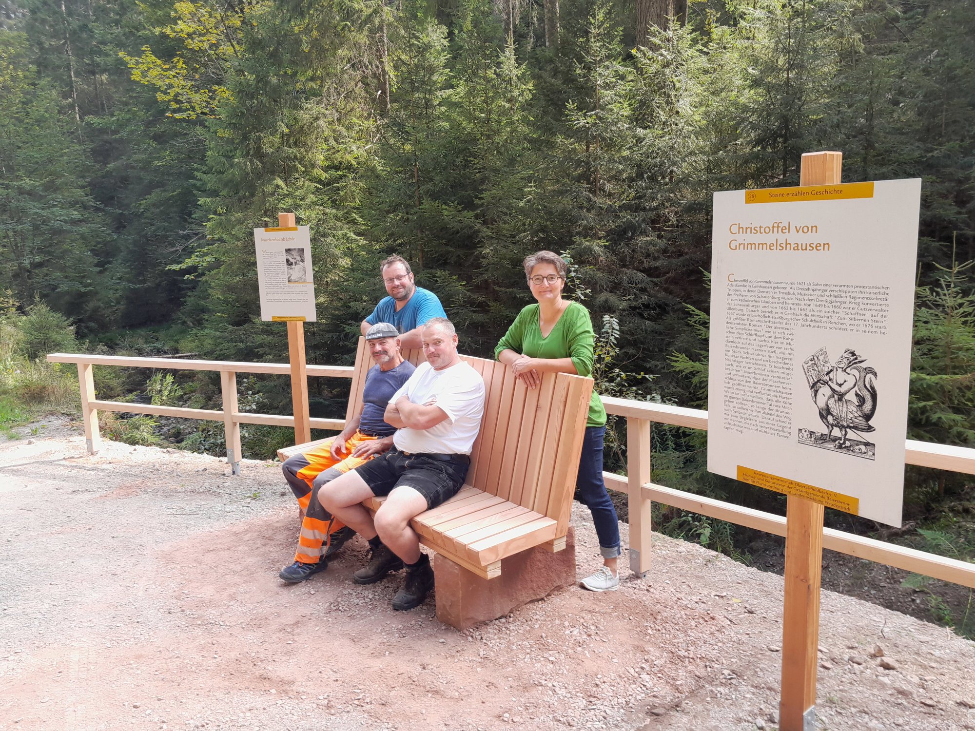 Neuer Aufenthaltsplatz am Muckenloch. © Dieter Dreher (Nationalpark Schwarzwald) Auf einem gschotterten Wegrand steht eine Holzbank, links und rechts davon je eine weiße Infotafel mit Bild und Text an einem hohen Holzpfahl. Auf der BAnk sitzen zwei Männer in Arbeitskleidung, hinter der Bank stehen noch ein Mann und eine Frau und schauen in die Kamera.
