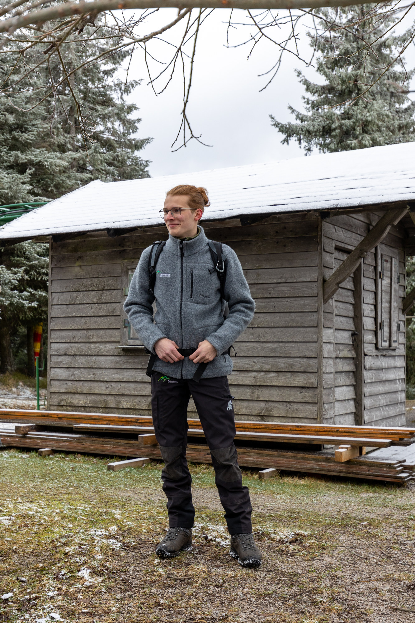 Ein junger Mann steht in grün-brainer Outdoorkleidung vor einer Waldhütte.