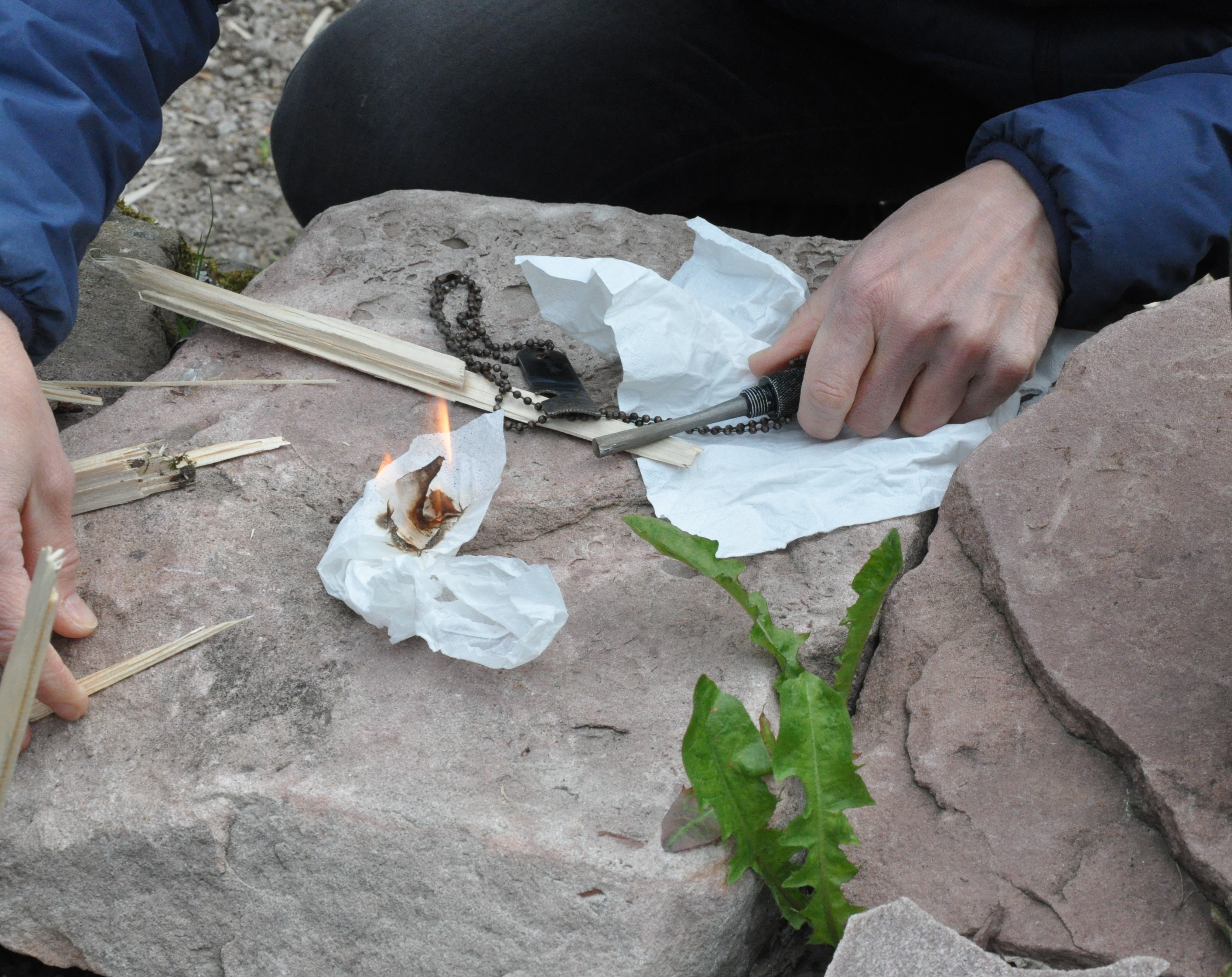 Zwei Hände versuchen mit Holz, Papier und Schlagstahl Feuer zu machen