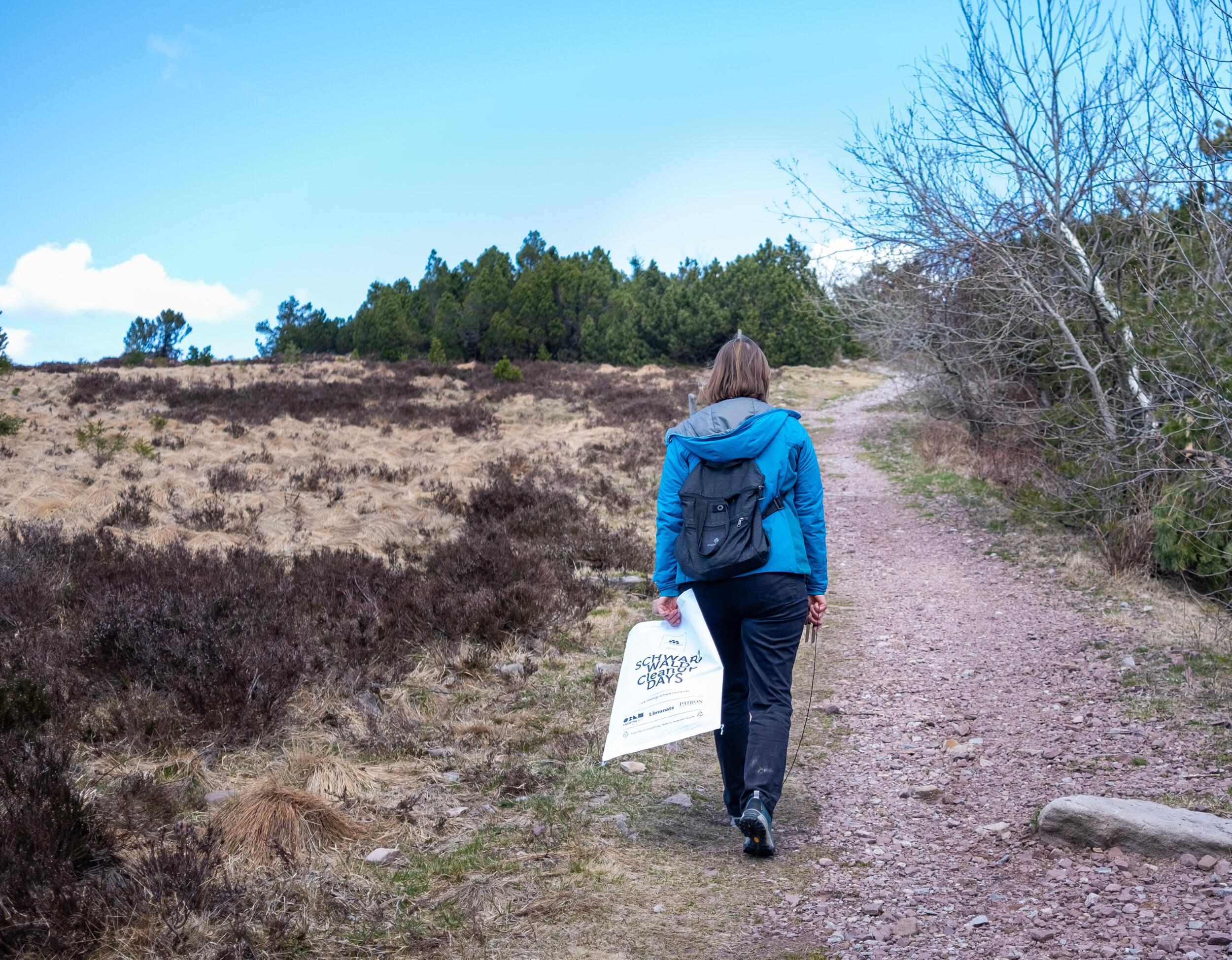 Frau in Wanderkleidung geht einen geschotterten Waldweg entlang. Sie hat eine Tüte in der Hand mit dem Aufdruck "Schwarzwald Cleanup Days"