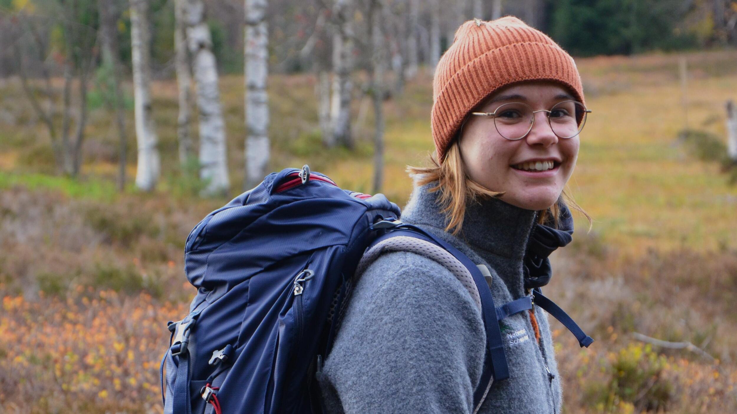 Die Freiwilligendienstleistende Elsa steht in der Natur vor einer Moorlandschaft mit Birken im Hintergrund und trägt eine Mütze