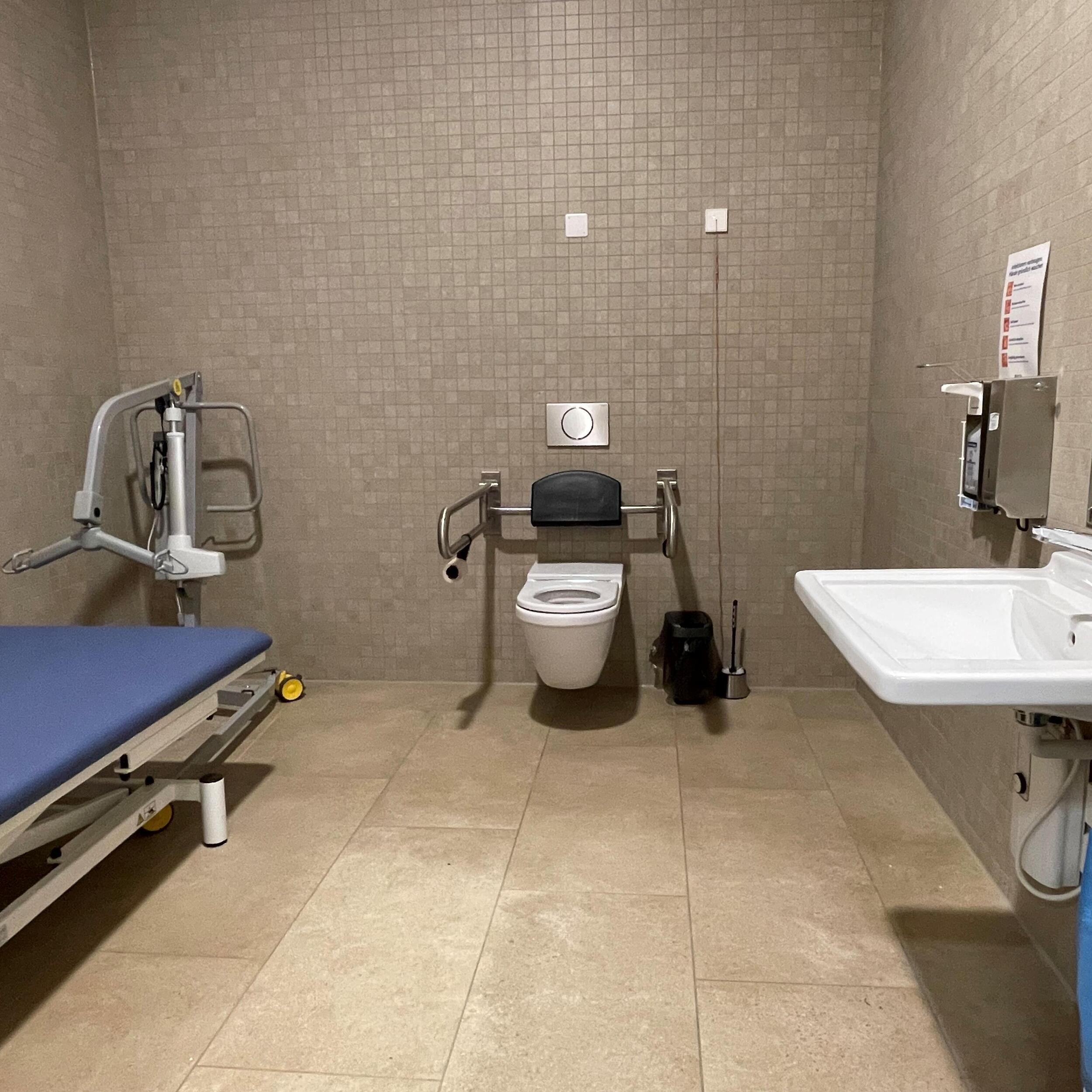Innenraum WC, Toilette mit seitlichen Stützen, höhenverstellbarer Liege und mobilem Lifter, sowie Waschbecken (nicht höhenverstellbar).