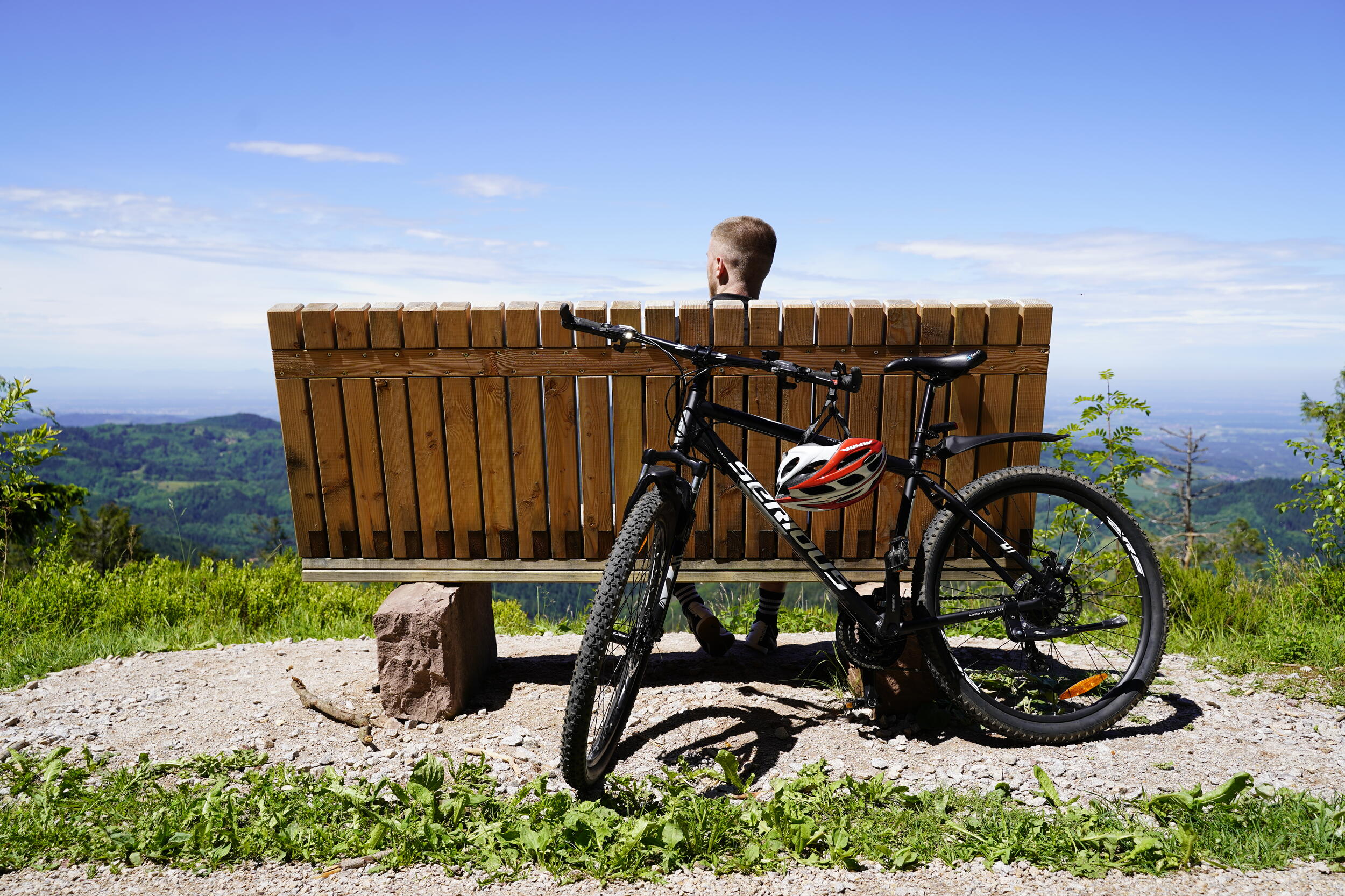 Ein Mountainbike lehnt an einer Holzbank, die von hinten zu sehen ist. Auf der Bank sitzt eine Person mit kurzen Haaren, nur der Hinterkopf ist sichtbar, die ins Tal schaut.