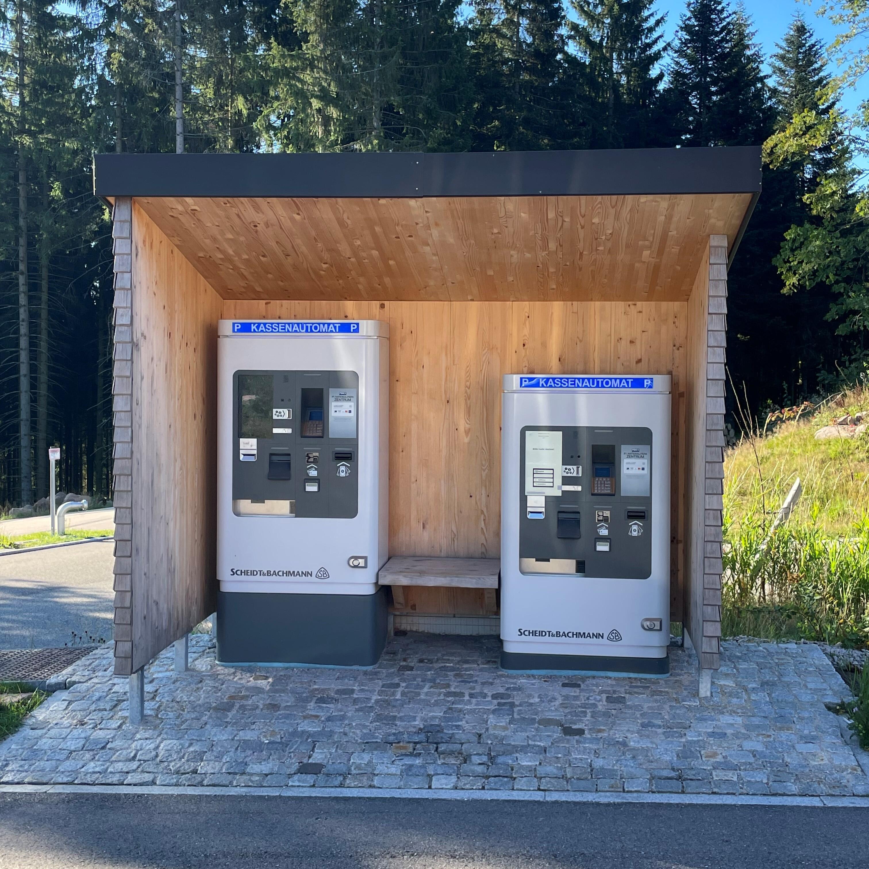 Frontalansicht Unterstand mit 2 Bezahlautomaten in unterschiedlicher Bedienhöhe. Davor Kopfsteinpflaster mit leichtem Anstieg. 