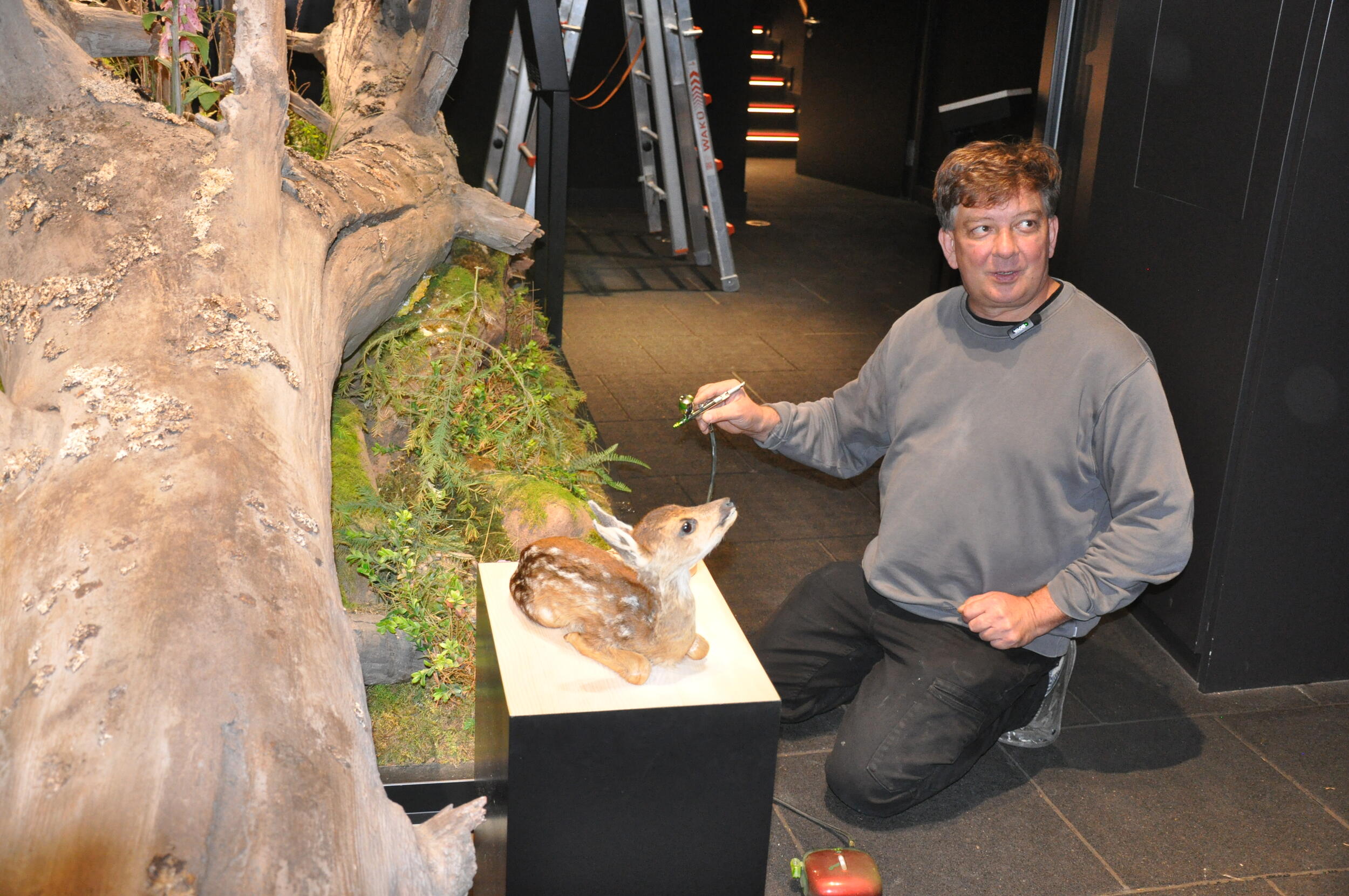 Ein Mann sitzt auf dem Boden neben einem künstlichen Rehkitz auf einem Podest. Er hält ein Instrument mit Farbe in der Hand. Foto: Iris Lemanczyk