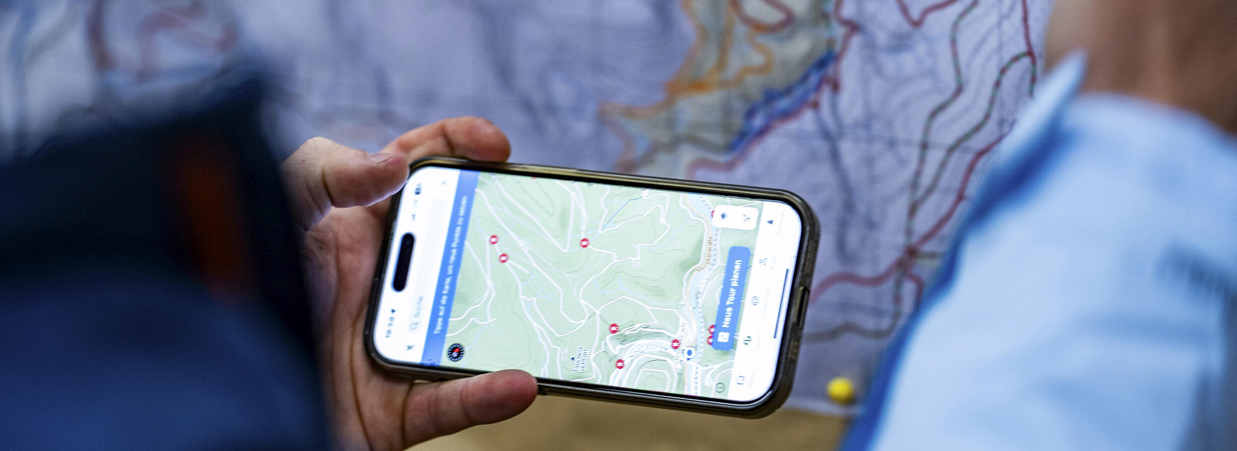 Eine Hand hält ein Handy mit einer Kartenansicht, im Hintergrund eine weitere Karte. Foto: Daniel Müller