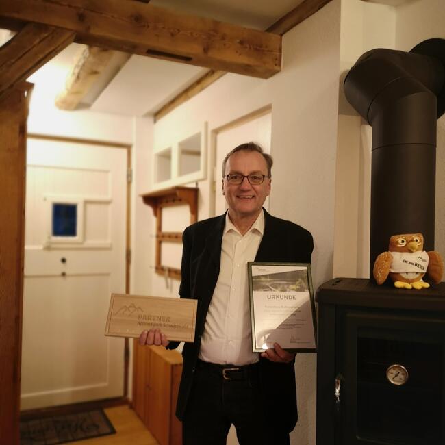 Ein Mann steht in einer Wohnung mit Ofen und hält in er einen Hand eine Urkunde, in der anderen ein Schild mit der Aufschrift Nationalpark Schwarzwald. Foto: Olivia Gerspach