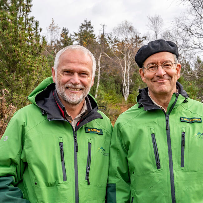 Zwei ältere Männer in grünen Outdoorjacken lachen in die Kamera.